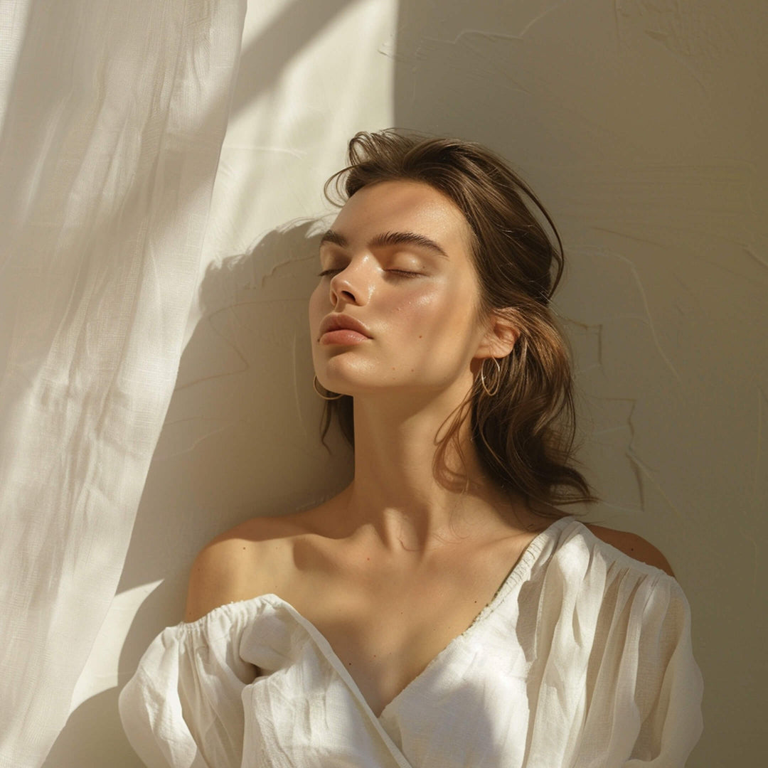 Woman in soft sunlight wearing an off-the-shoulder white blouse, eyes closed and face illuminated by warm natural light, evoking calm and radiant skin.