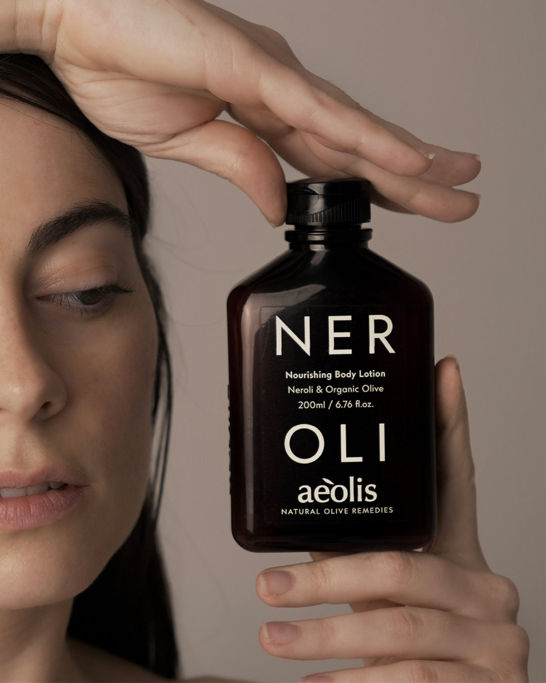 Bottle of Aeolis Neroli Nourishing Body Lotion held by a hand with a woman's face in the background.