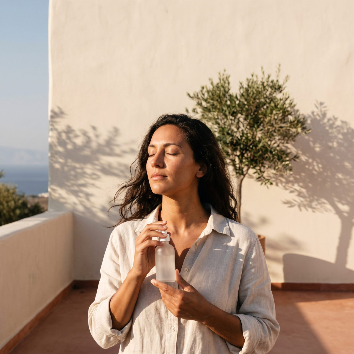 Woman in a linen shirt, eyes closed in afternoon sun on a whitewashed Mediterranean terrace, an olive tree and the sea behind her